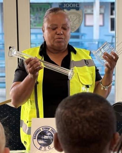 A woman wearing a yellow safety vest is holding a rain gauge while speaking to a group of children. A sign that says 