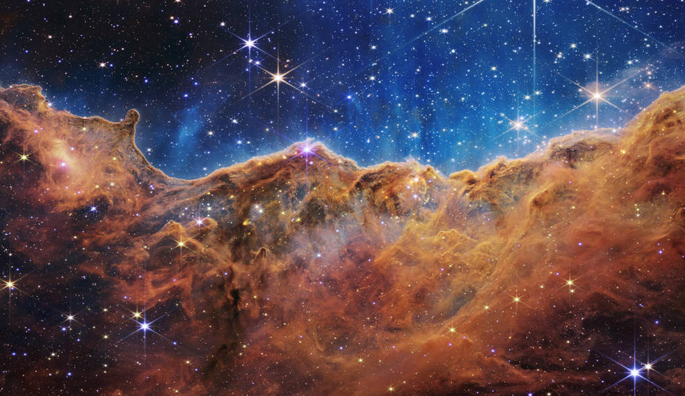 An undulating, translucent star-forming region in the Carina Nebula is shown in this Webb image, hued in ambers and blues; foreground stars with diffraction spikes can be seen, as can a speckling of background points of light through the cloudy nebula.