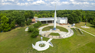 An afternoon view above the rocket garden at NASA Goddard's Visitor Center. The prominent Delta B launch vehicle stands 90-feet tall and was a type used to orbit satellites in the early 1960s. In the foreground, a kinetic sculpture called Orbits Interweave incorporates three polished stainless steel spheres — representing the Sun, Earth, and GOES weather satellites — that move gently in the wind. The exhibit area around it is shaped like a hurricane symbol. Imaged June 13, 2023, looking east-southeast.Credit: NASA/Francis Reddy