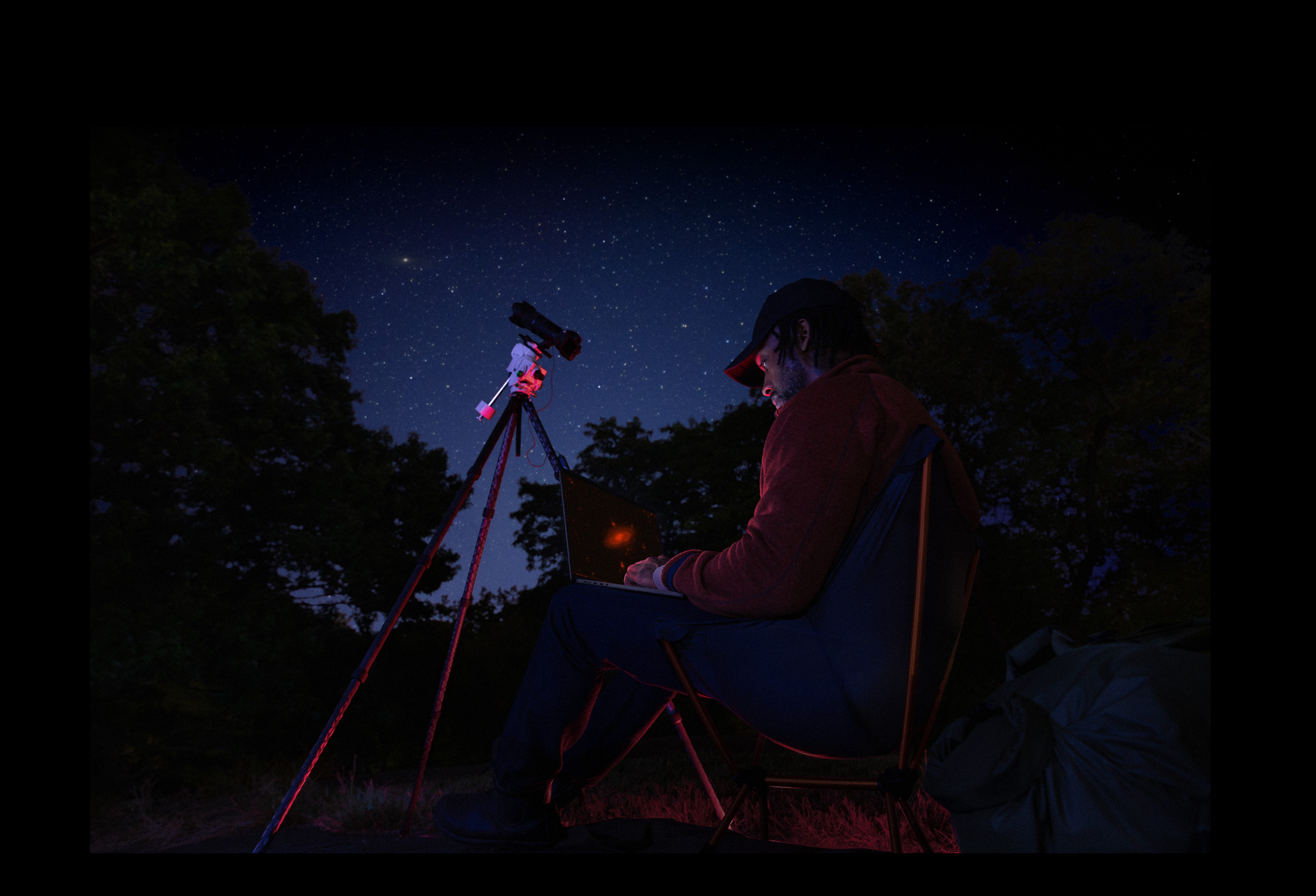 Dehors en pleine nuit, une personne tient sur ses genoux un MacBook Pro connecté à un appareil installé sur un trépied et pointé vers le ciel étoilé