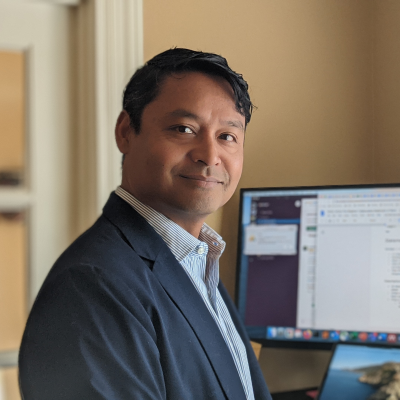 The is a rectangular, landscaped-oriented portrait of Dr. Rahul Ramachandran in a office. Ramachandran is standing on the left side of the image in front of a desk and computer monitor with a window on the right. Ramachandran is wearing a dark sport coat with a light-colored collared shirt.