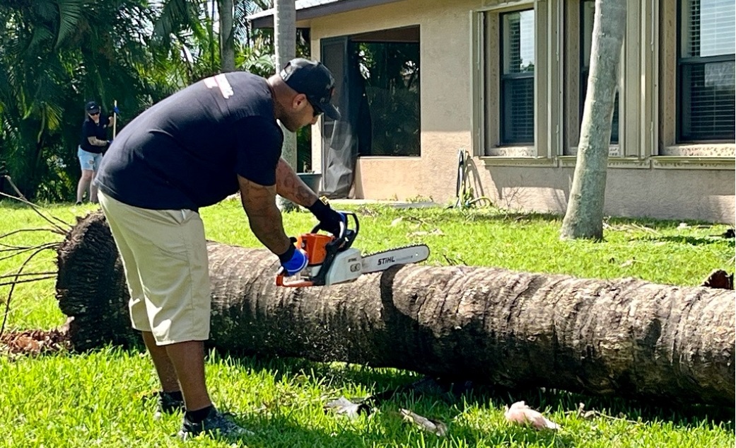 A volunteer using a chainsaw to cut up a fallen tree in front of a house.