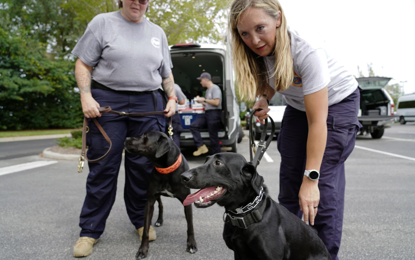 Two women holding two black dogs standing in a parking lot