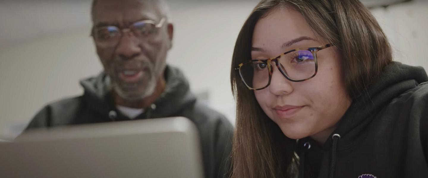 A young girl stares into the screen of a computer laptop while her teacher looks on in the background.