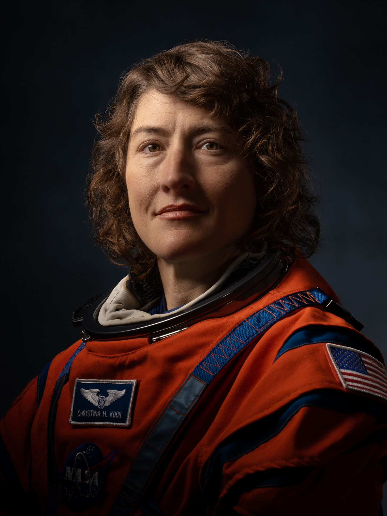 NASA astronaut Christina Hammock Koch poses for a portrait in her orange Artemis flight suit. The suit has blue trim around the neck and shoulders, with three patches: one with the U.S. flag on her left shoulder, one with her name and a pair of wings on her chest, and one with the NASA “meatball” insignia faintly visible beneath the second. The background is dark, and the photo is lit to focus on Koch’s face, which is facing the camera with a dignified expression. Credit: NASA