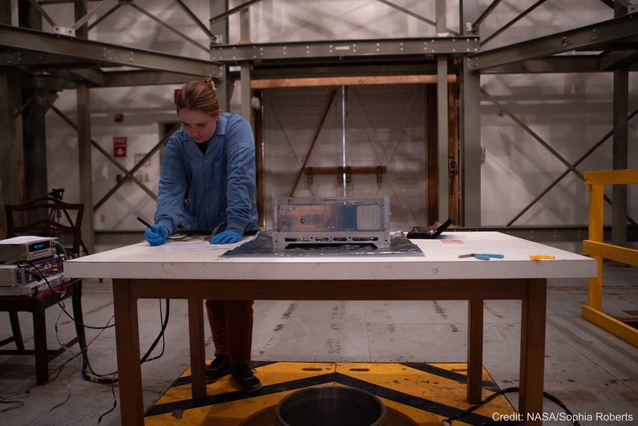 This image shows a woman wearing a long-sleeved blue jacket and blue gloves. Her hair is bound up in a clip. She leans over a table, filling out a form. To the right, on the same table, is a shiny box within another clear box — the BurstCube satellite in its protective case. The dim room behind the woman is full of gray beams that cast shadows against the walls. There is an old white barn door in the far wall. The image is watermarked, “Credit: NASA/Sophia Roberts.”