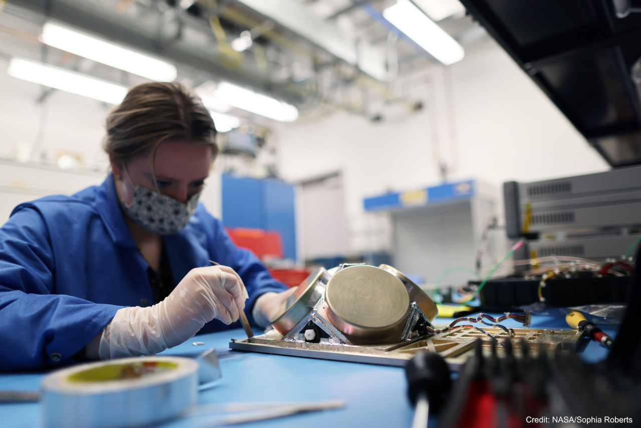 Julie Cox, a mechanical engineer at Goddard, presses aluminized tape to the BurstCube instrument in a laboratory. Julie is wearing a mask, blue lab coat, and gloves, and is holding silver tweezers in one hand. The instrument, which is sitting on a table covered in hardware and tools, has raised silver-colored metal cylinders on top of a flat plate with triangular and rectangular cutouts. A roll of tape sits on the table in the foreground. The image is watermarked with “Credit: NASA/Sophia Roberts.”