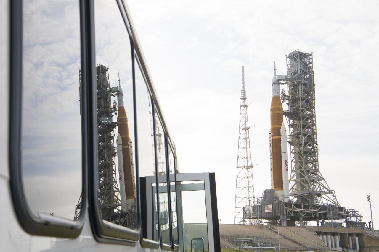 Cloudy skies are the backdrop behind the SLS rocket and Orion spacecraft, which is reflected in the windows of a vehicle to the left of the photo. The SLS is orange with two white boosters on either side, and the spacecraft is white, next to a gray pyramid-shaped lightning tower and Mobile Launcher with many pipes and levels in gray and red. Credit: NASA/Aubrey Gemignani