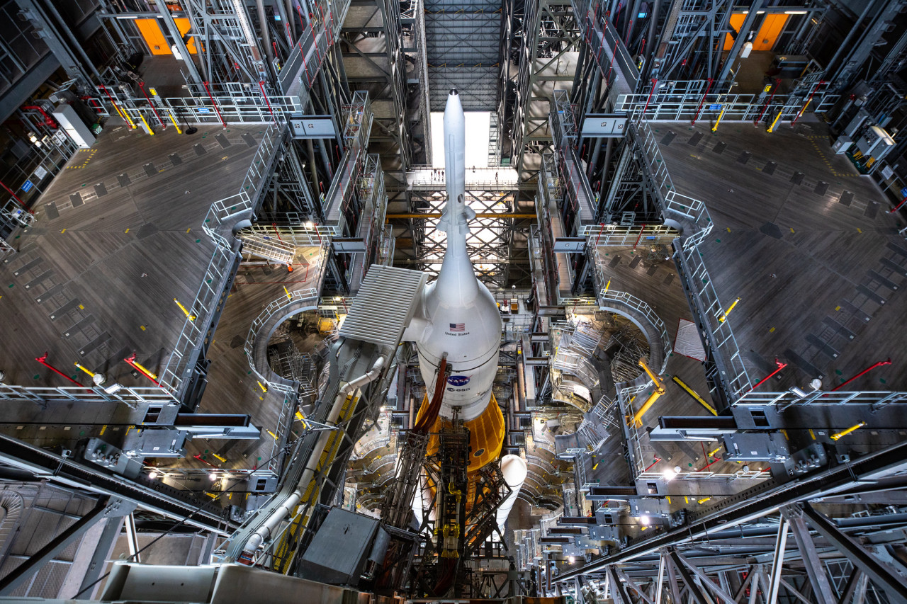 The SLS rocket and Orion spacecraft sit inside the Vehicle Assembly Building (VAB) at Kennedy Space Center. The rocket is orange, with two white boosters on either side. The Orion Spacecraft is at the top and white. The VAB has many levels with walkways, pipes, and structures around the rocket. Credit: NASA/Kim Shiflett