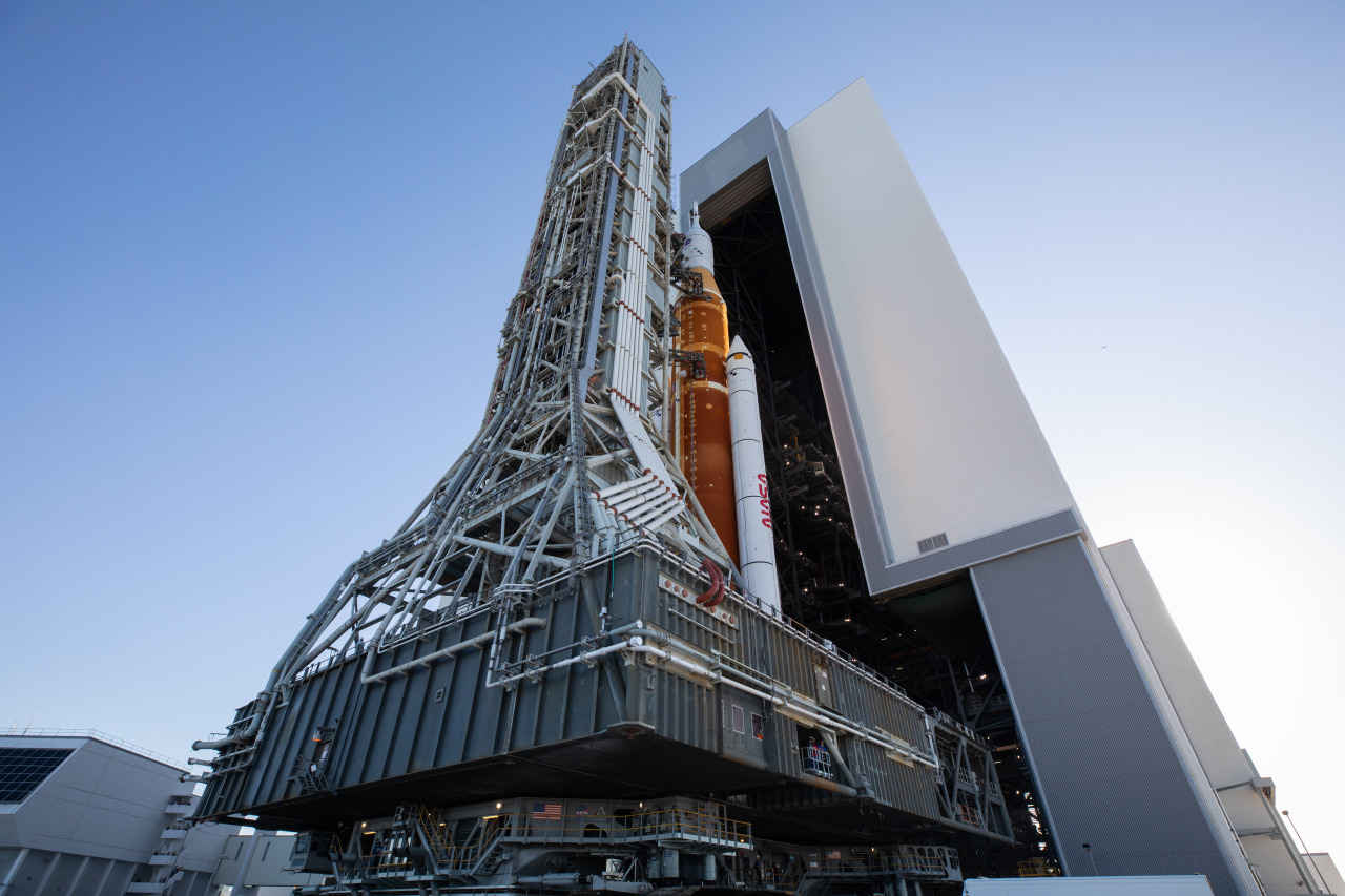 NASA’s Space Launch System (SLS) rocket, with the Orion capsule atop, slowly rolls out of the Vehicle Assembly Building on the crawler-transporter 2. The crawler is grey with treads and walkways, and the SLS is orange with two white boosters on either side.  Credit: NASA/Kim Shiflett