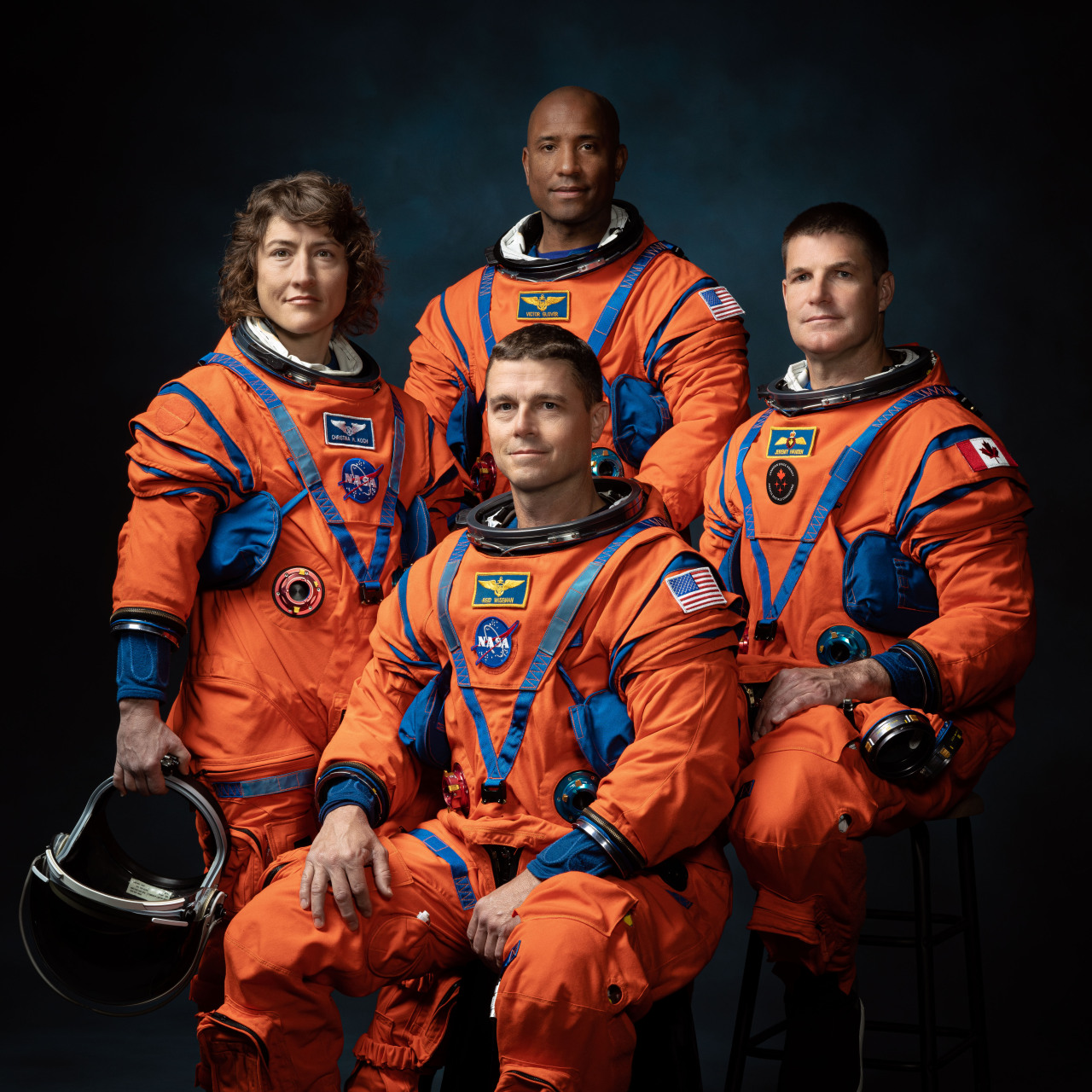 The Artemis II crew sits for an official portrait in front of a dark background. They wear orange suits with various patches noting their names, nationalities, and NASA or CSA. From left to right, are NASA astronauts Christina Koch, Victor Glover (top), and Reid Wiseman (bottom), and Canadian Space Agency astronaut Jeremy Hansen. Koch holds a helmet in her hand. Credit: NASA