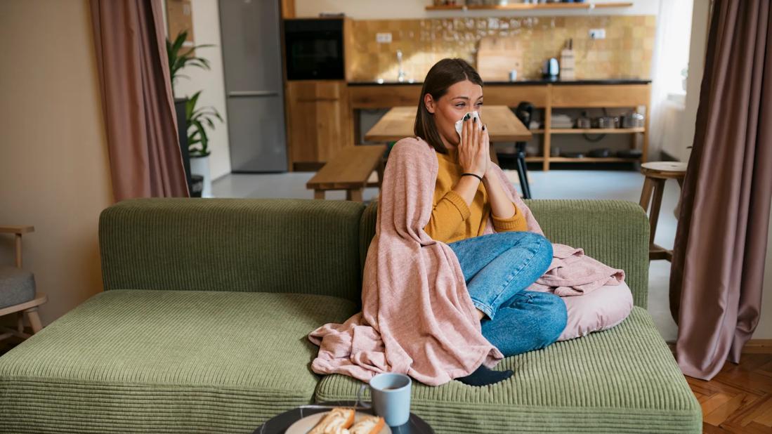 Person sitting on couch at home, wrapped in blanket, blowing their nose, with toast and tea on table