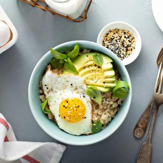 Bowl of oatmeal, topped with fried egg and avocado, with small bowl of seeds on table