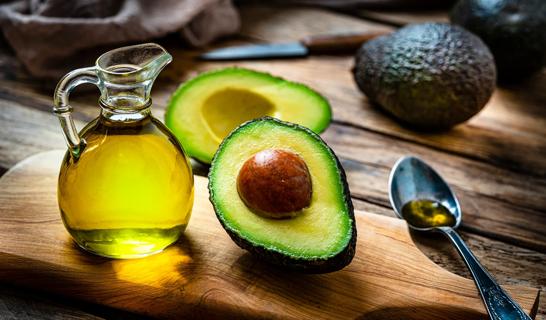 Half an avocado with the pit inside sits next to a glass pitcher of avocado oil and a spoon with some oil on it