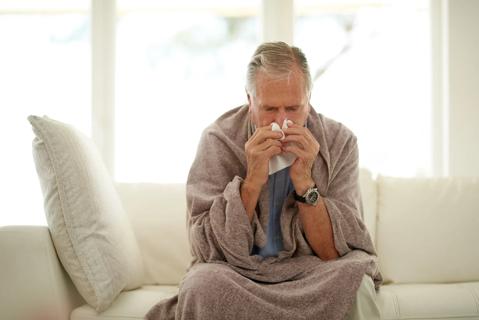 Older person sitting on couch, wrapped in blanket, hunched over, blowing their nose