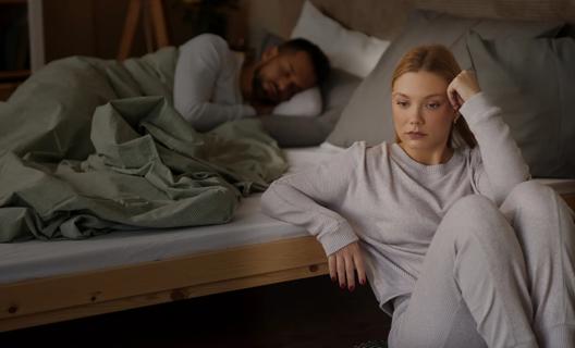 Person sitting on floor at night next to bed in deep thought, with partner sleeping in bed