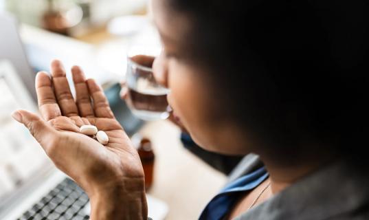 Person holding two pills in palm of hand and glass of water in other hand, keyboard in background