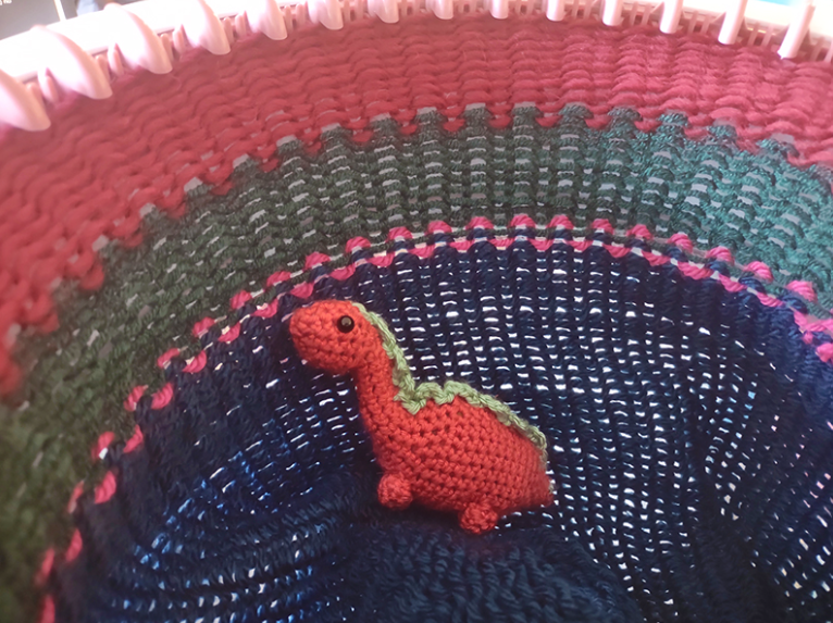 A mini, crocheted red brontosaurus with green spikes sits in the center of a round knitting machine, on the material already knit in stripes of red, green, pink, and blue.