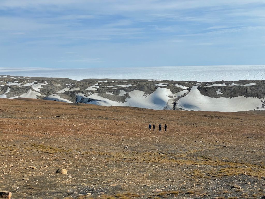 The Greenland landscape. The lower part of the image shows brown rocky ground with four tiny dots of people walking toward the ice sheet in the midground. The horizon is above the ice show blue sky lightly overcast with a sheet of white bumpy clouds.