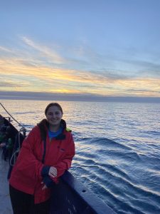 Mackenzie, a young white woman in a long red coat, poses on the R/V Bold Horizon. She is leaning on the railing, with blue ocean water and a sunset behind her.