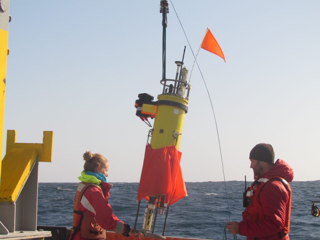 Two people stand to either side of a about five-foot tall cylindrical instrument they are prepareing to put in the water.