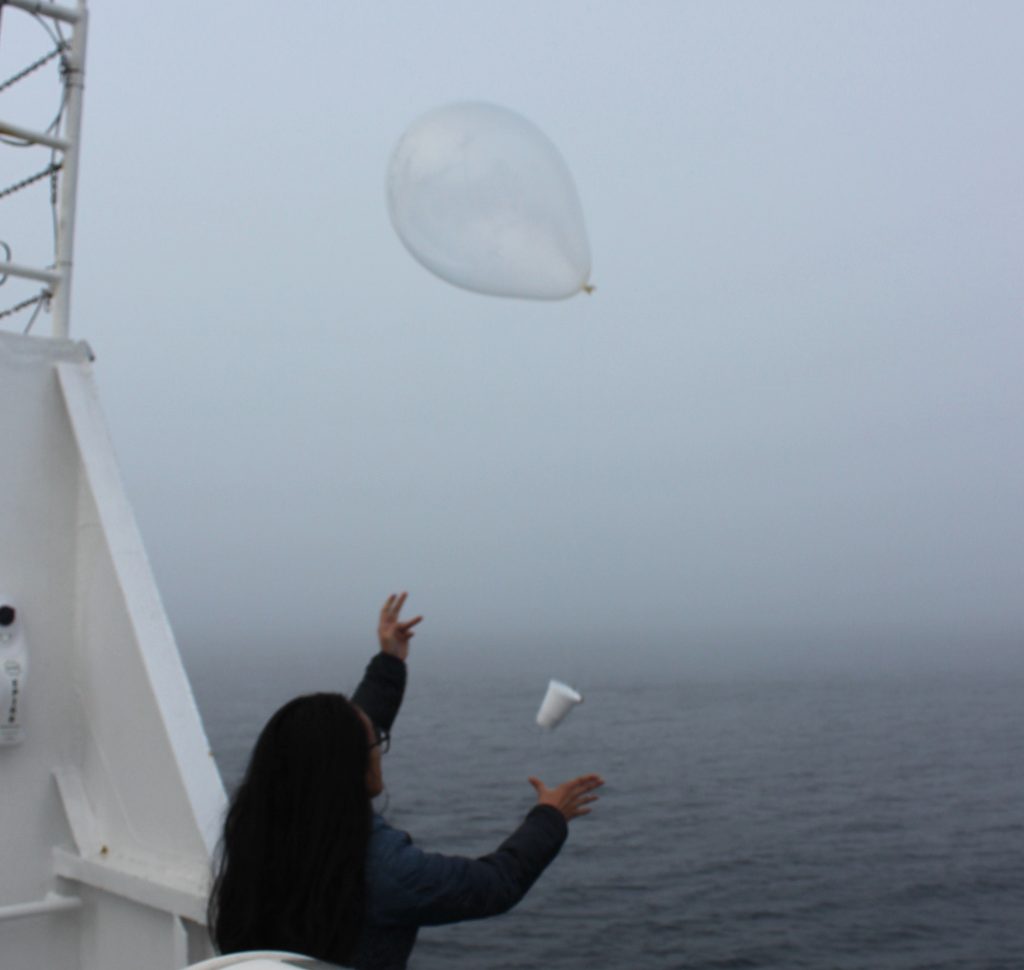 A woman stands at the rail of a boat, ocean in the background. Her hands are up as they have just let go of a balloon with a white instrument the size of a cup dangling from it.