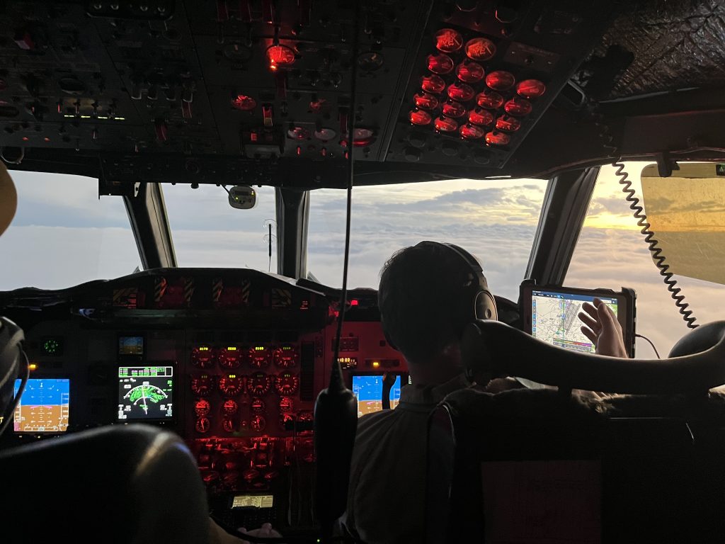 Photo into the cockpit of a plane, flying over storm clouds around sunset. There are two pilots in the cockpit, each with their own steering devices and monitors and gauges.