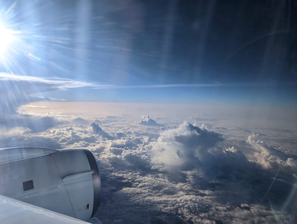 Photo taken out of a plane window. Part of the engine is seen in the lower left corner. The sky is blue, but the lower part is clouded with puffy clouds and brown dust.