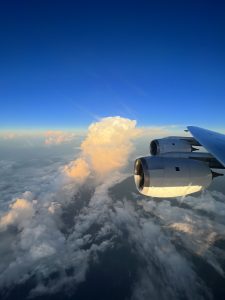 Photo taken out of a plane window. Part of the engine is seen on the right side. The sky is blue, but the lower part is clouded with puffy clouds and brown dust.