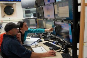 Two researchers sit in front of 12 computer screens, reading a display of data. The computer monitors are aboard a research vessel, with a port hole window in the background.