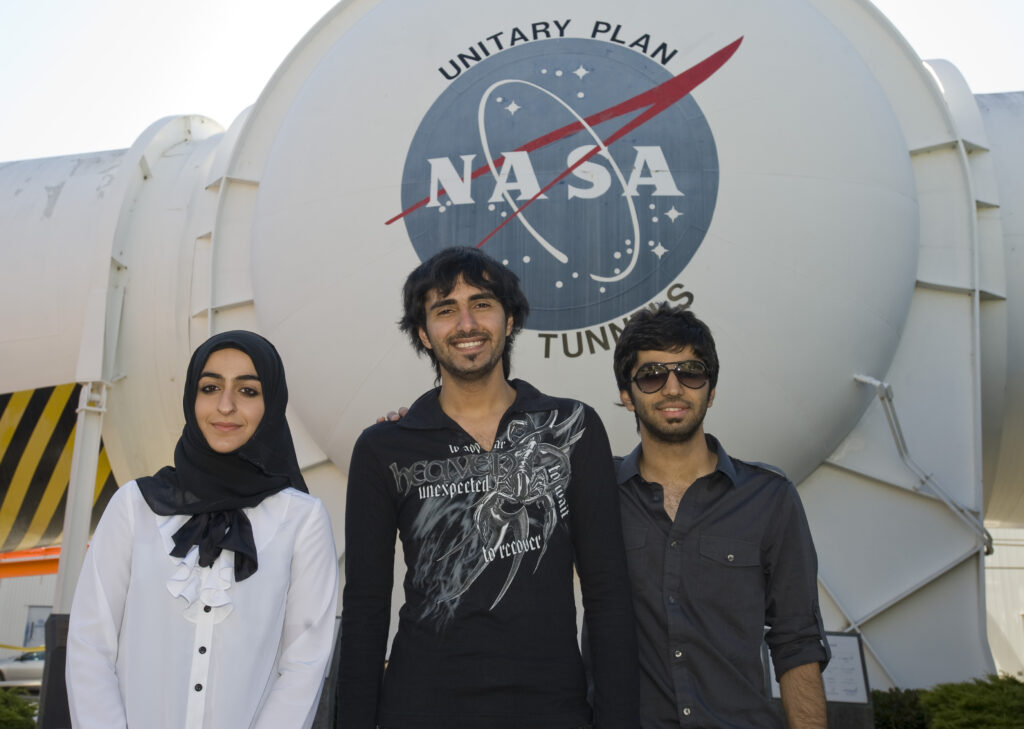 Three international NASA interns from the United Arab Emirates pose together outside of the large Unitary Plan Wind Tunnel, located at the NASA Ames Research Center. Wearing business casual attire, the trio smile in front of the massive white building housing the wind tunnel. Credit: NASA / Eric James