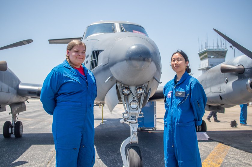 Alt Text: Two female interns wearing blue NASA flight suits stand smiling with their hands behind their backs. Behind them, a large multirotor aircraft sits idle on the runway. The distance clear blue sky makes up the top of the image. Credit: NASA/Angelique Herring