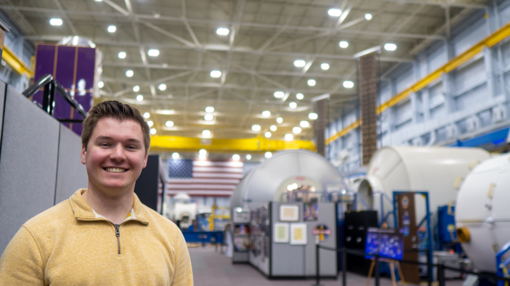 Alt Text: Matthew McDonnell stands in a large hangar at NASA's Johnson Space Center. He wears a light-colored quarter-zip shirt and looks at the camera. In the background a large American flag hangs on the wall, the metallic structure of the hangar illuminated by large overhead lights. Near Matthew large rounded space capsules are visible. Credit: Matthew McDonnell