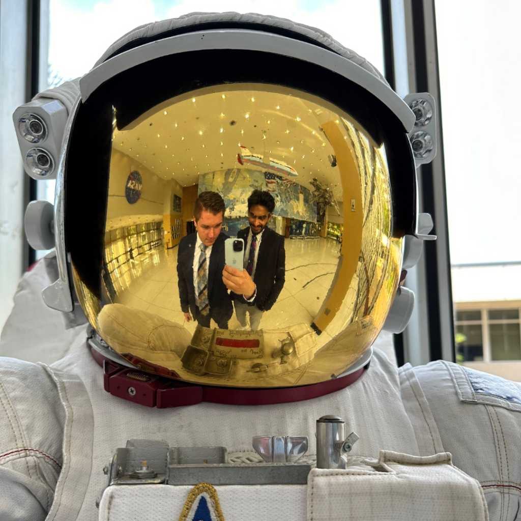 Two NASA interns take a selfie together in the reflective visor of a spacesuit. The yellow hues of the visor tints the reflected image. 