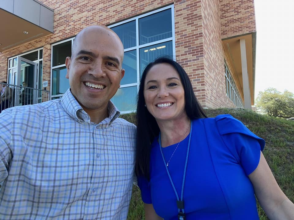 Alt-text: NASA’s Johnson Space Center SkillBridge Coordinator, Mr. Albert Meza wearing a plaid dress shirt and Trish wearing a blue dress with a lanyard. The two are smiling and pictured left from right in a selfie in front of a brick building.