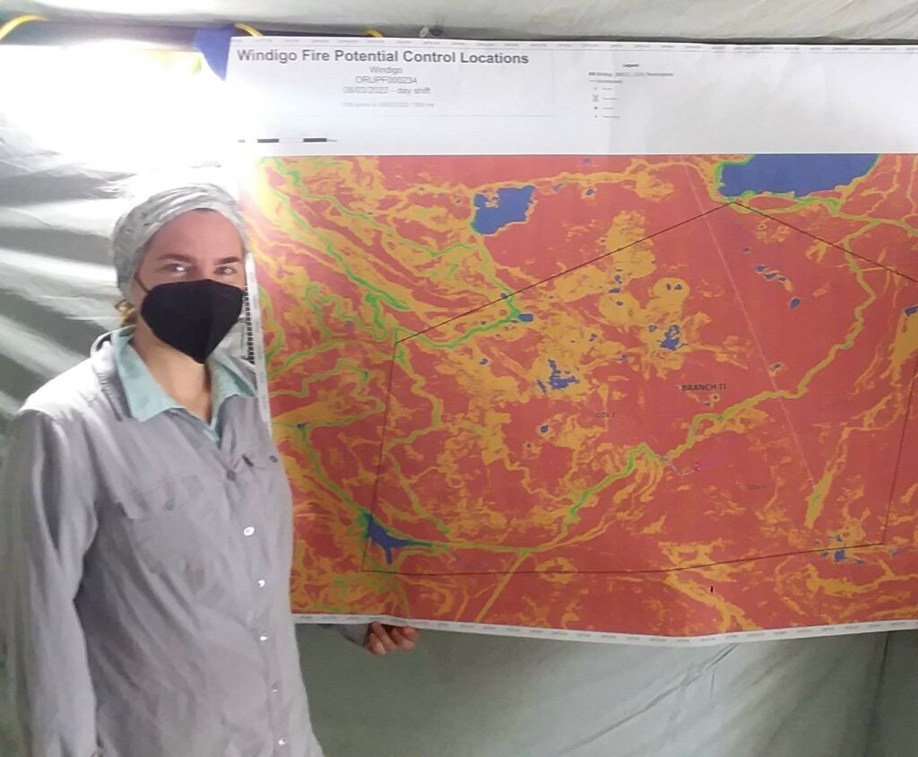 Alt Text: Jenessa Stemke poses next to a map of potential fire control locations. She is wearing a gray shirt, face mask and headband. Credit: Bryce Foster