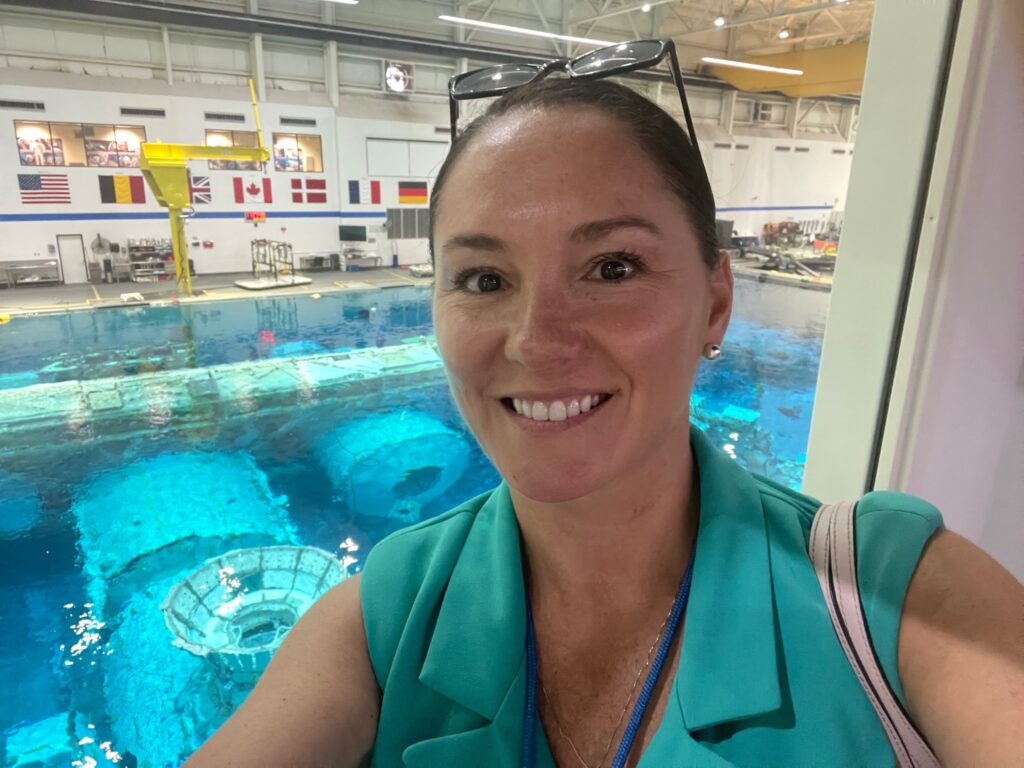 Former NASA skill bridge intern Trish Elliston take a selfie in NASA's Neutral Buoyancy Laboratory. A large pool of water is visible in the background inside a massive hangar like building. Trish smiles at the camera wearing a light blue top with her hair pulled back. Credit: Trish Elliston 