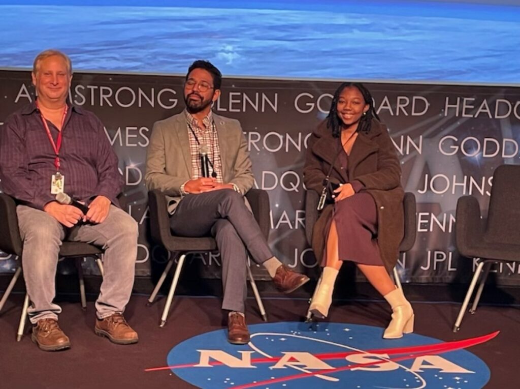 NASA intern Asia sits beside two NASA employees in professional attire at a conference. Below them, the carpet brandishes a NASA meatball logo. In addition, we see the names of NASA centers obscured behind them.