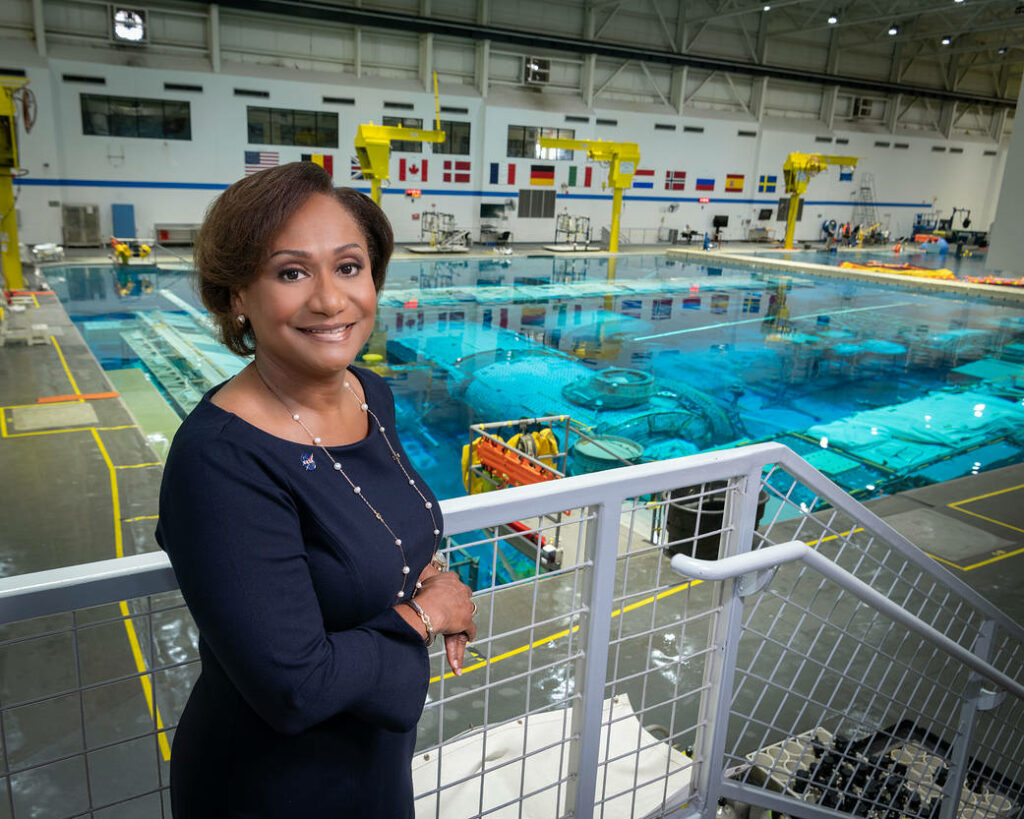 Alt Text; NASA's Johnson Space Center Director Vanessa Wyche poses before the Neutral Buoyancy Lab. Wearing a dress, Wyche smiles in front of the large pool which astronauts train within. In the background a large facility includes hanging flags of member nations of the International Space Station. Image Credit: NASA/Bill Stafford