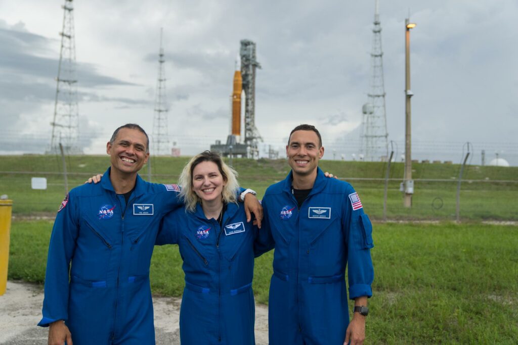 Alt Text: NASA astronaut candidates Anil Menon, Deniz Burnham, and Marcos Berrios pose for a photograph in front of NASA’s Artemis I Space Launch System and Orion spacecraft atop the mobile launcher on the pad at Launch Complex 39B. The trio wear blue flight suits and smile. In the background the orange structure of the SLS towers open grass fields. Image Credit: NASA/Steven Seipel