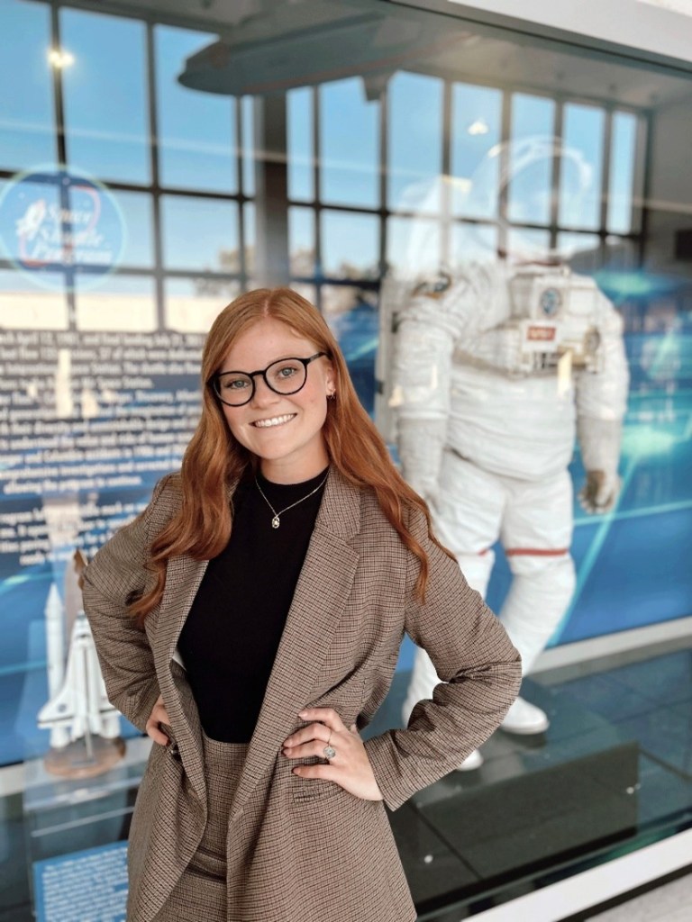 Alt Text: Mackenzi Haub, white woman with red hair and blue eyes, smiles while standing with her hands on her hips. Haub wears a brown suit with a black shirt. Behind her is a NASA display case for the Space Shuttle Program and an astronaut suit. 