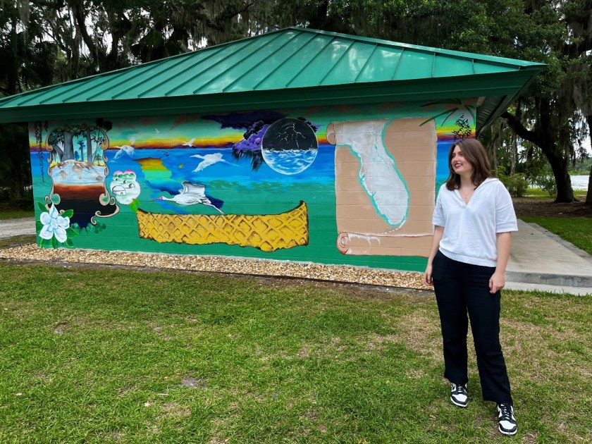 Alt text: Berlie stands to the right of a mural in progress, which she led as part of her environmental fellowship. The mural depicts an aerial bird’s-eye view of where Lake Beresford meets the St. John’s River, references the river’s native projectile points, shell mounds, snails, pottery designs, and includes to the far right a scroll with the state of Florida. Berlie wears black and white sneakers, black pants, and a white V-neck shirt.