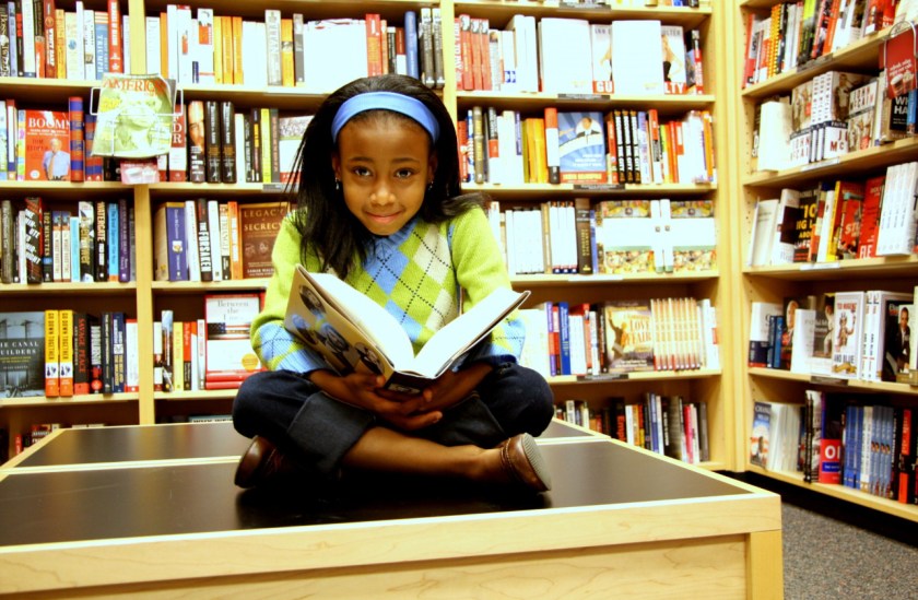 Alt Text: NASA intern Caitlyn McClanahan as a young student sitting on a table in a book store smiling with a large history book open upon her lap. Credit: Caitlyn McClanahan