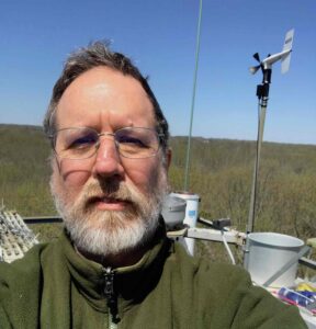 The image is a selfie of a bearded man seen from the shoulders up. He is wearing a dark green zip-up jacket, and also has glasses on. Behind him are scientific instruments, which look like long metal rods standing up as well as some buckets. In the far background is a grassy field and a clear blue sky.