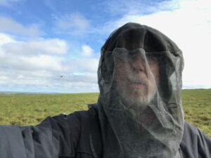 The image is a selfie of a man seen from the shoulders up. He is wearing a dark colored shirt and has a netted material covering his entire heat. His face can be seen through the netting and he is wearing glasses and has a beard. Behind him is a large grassy field. The sky is bright blue and has some white fluffy clouds.