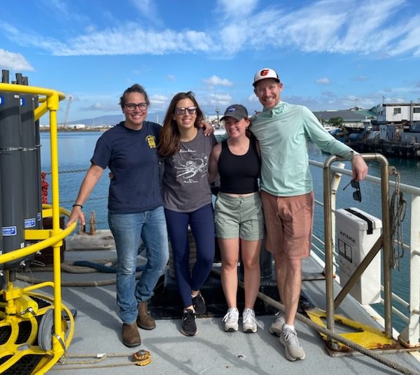 Four people stand on the deck of a boat with their arms around each other posing for the camera. From left to right: a woman is wearing a navy blue t-shirt and jeans, a woman is wearing a gray t-shirt and dark blue jeans, a woman is wearing a black tank top and teal shorts, a man is wearing a teal shirt and rust colored shorts. The two people on the outer edge are holding onto railings beside them. In the background of the image, the sky is bright blue with some small wispy clouds.