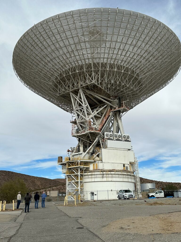 This is the 70 meter antenna at Goldstone, California.  The dish is enormous, taking up most of the image. The dish, which is white with lines running through it is turned up to the sky. It has a white base that attaches it to the ground.  Four people stand to the left giving a sense of scale.  Behind it are low mountains and a mostly cloudy sky. 