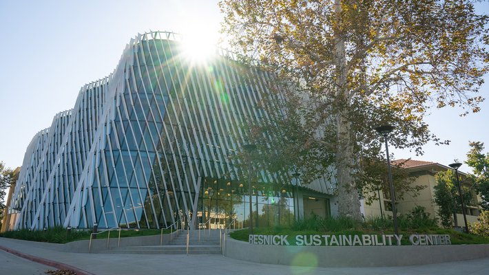 A building with a curved glass facade and diagonal shade fins on the exterior is shown, illuminated by the sun, with a sign reading Resnick Sustainability Center in silver letters in the foreground.