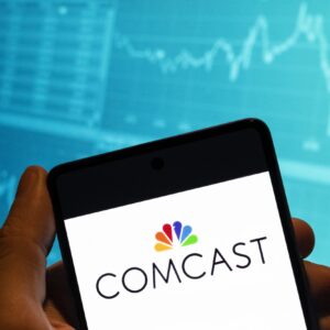 In this photo illustration, the Comcast logo is displayed on a smartphone with a stock exchange index graph in the background.