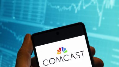 In this photo illustration, the Comcast logo is displayed on a smartphone with a stock exchange index graph in the background.